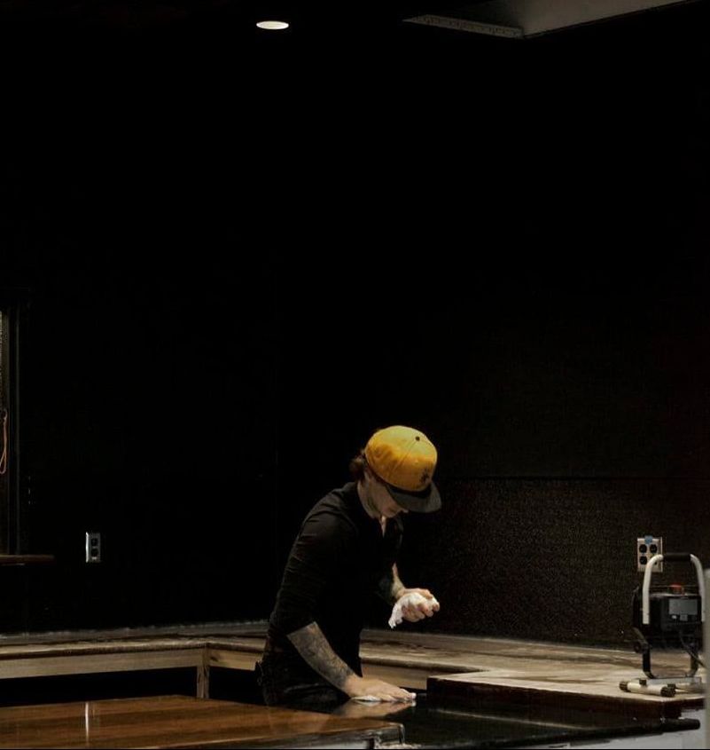 Person in a yellow hat, squatting, leaning over a wood surface, possibly preparing food. Black background.