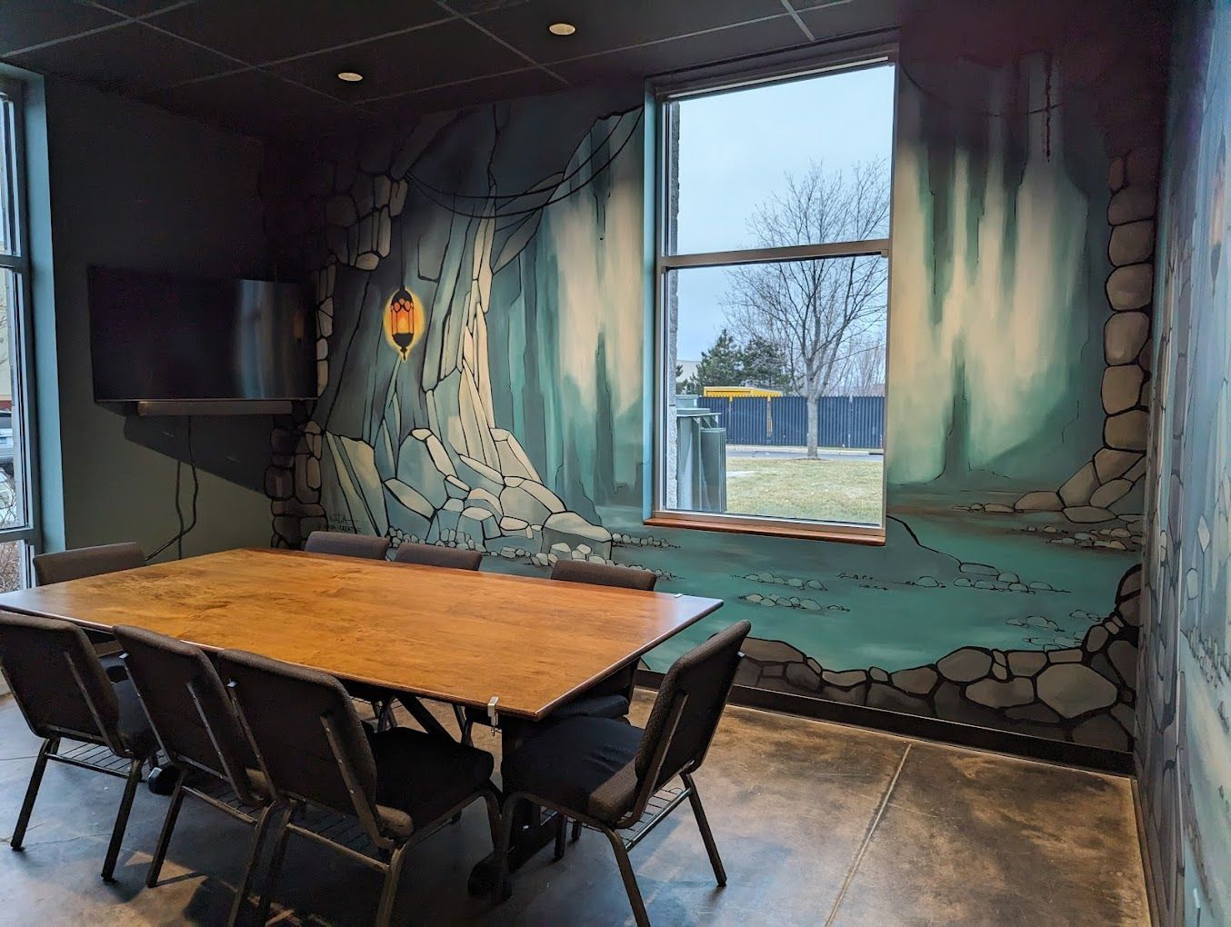A conference room with a wooden table, chairs, and forest mural. A window shows the outside.