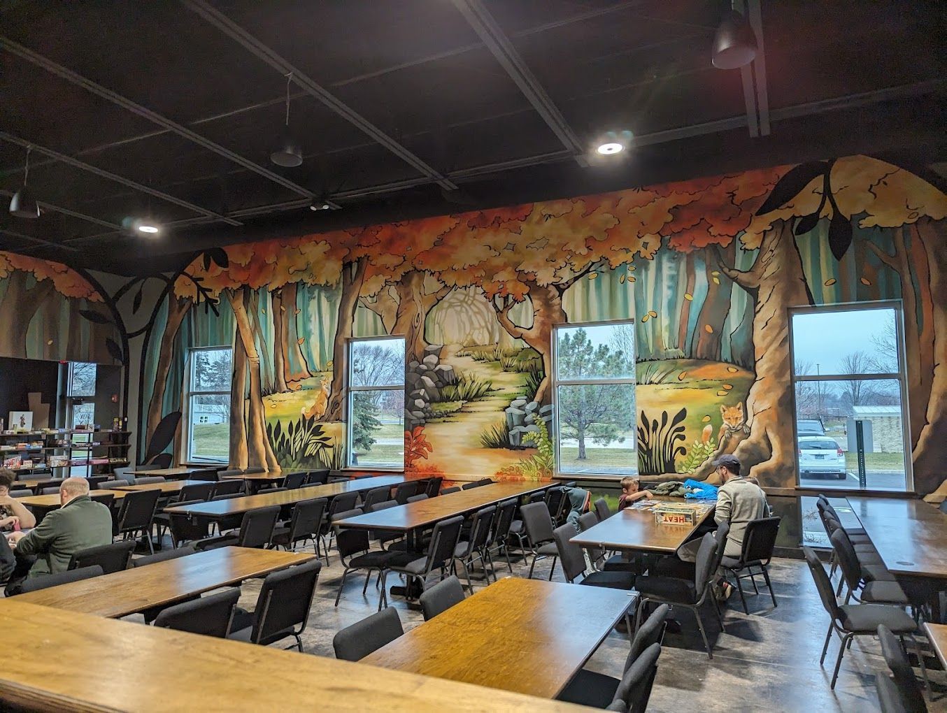 Indoor dining area with forest mural and long tables. People are seated. Windows offer outdoor view.