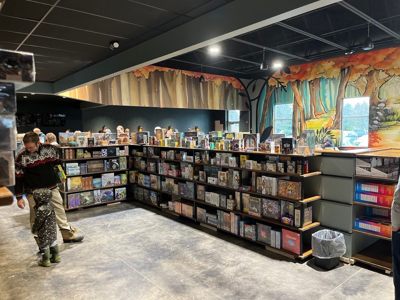 Board game store interior with shelves full of games, mural, people browsing.