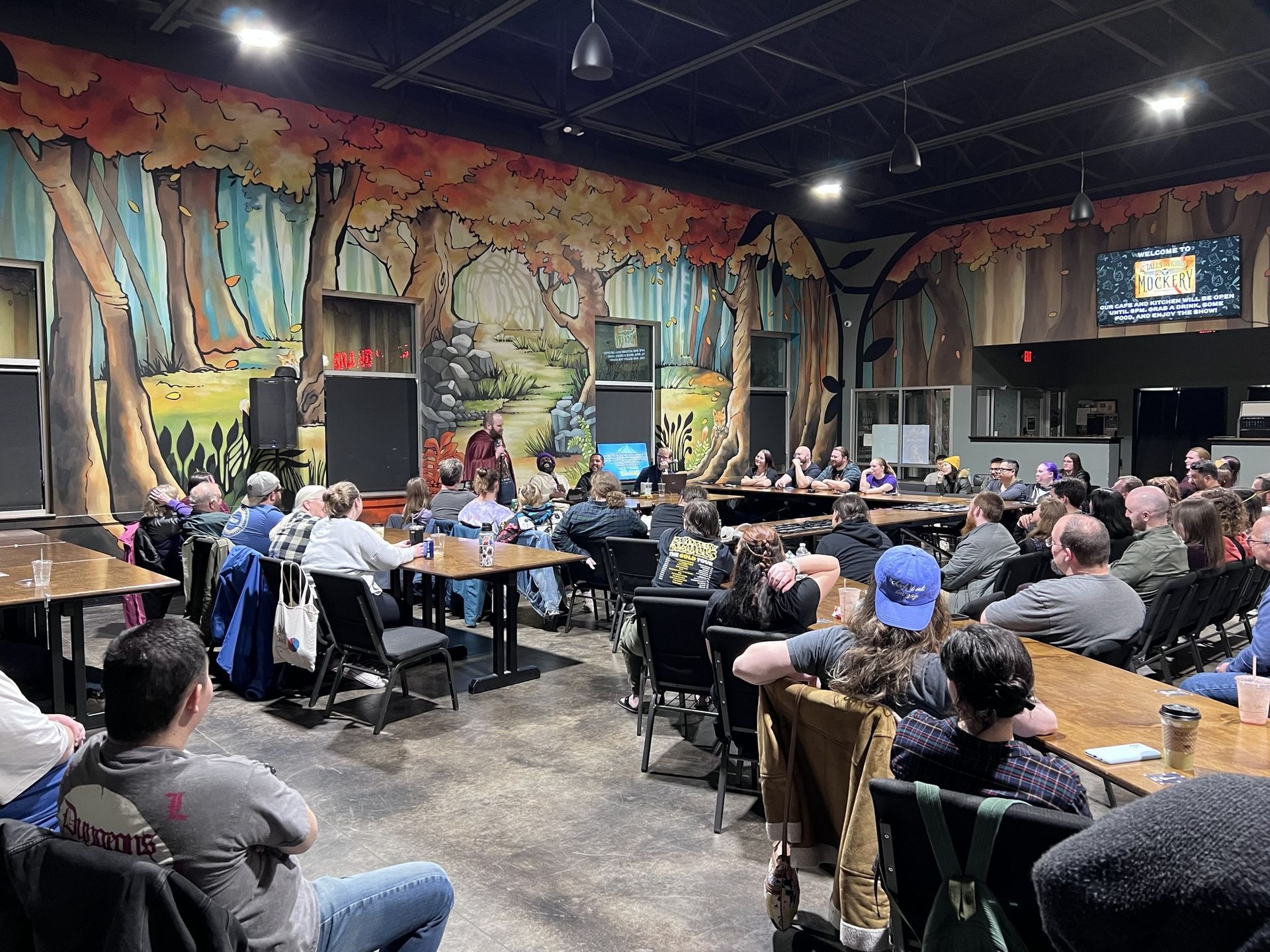 Meeting in a room with a mural. People sit at tables, listening to speakers at a table on a stage.