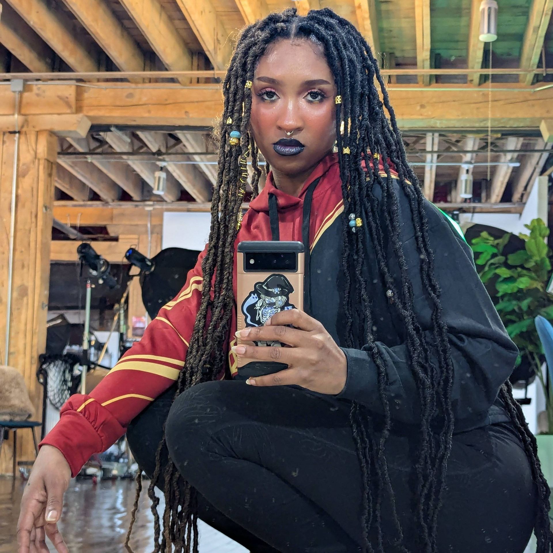 Person with locs, in a Harry Potter hoodie, holding phone. Dark makeup, squatting pose, indoor setting.