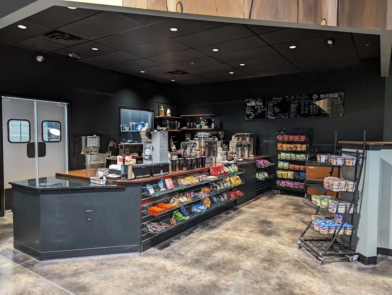 Interior view of a small cafe, black walls, counter with snacks, coffee machines, and shelves.