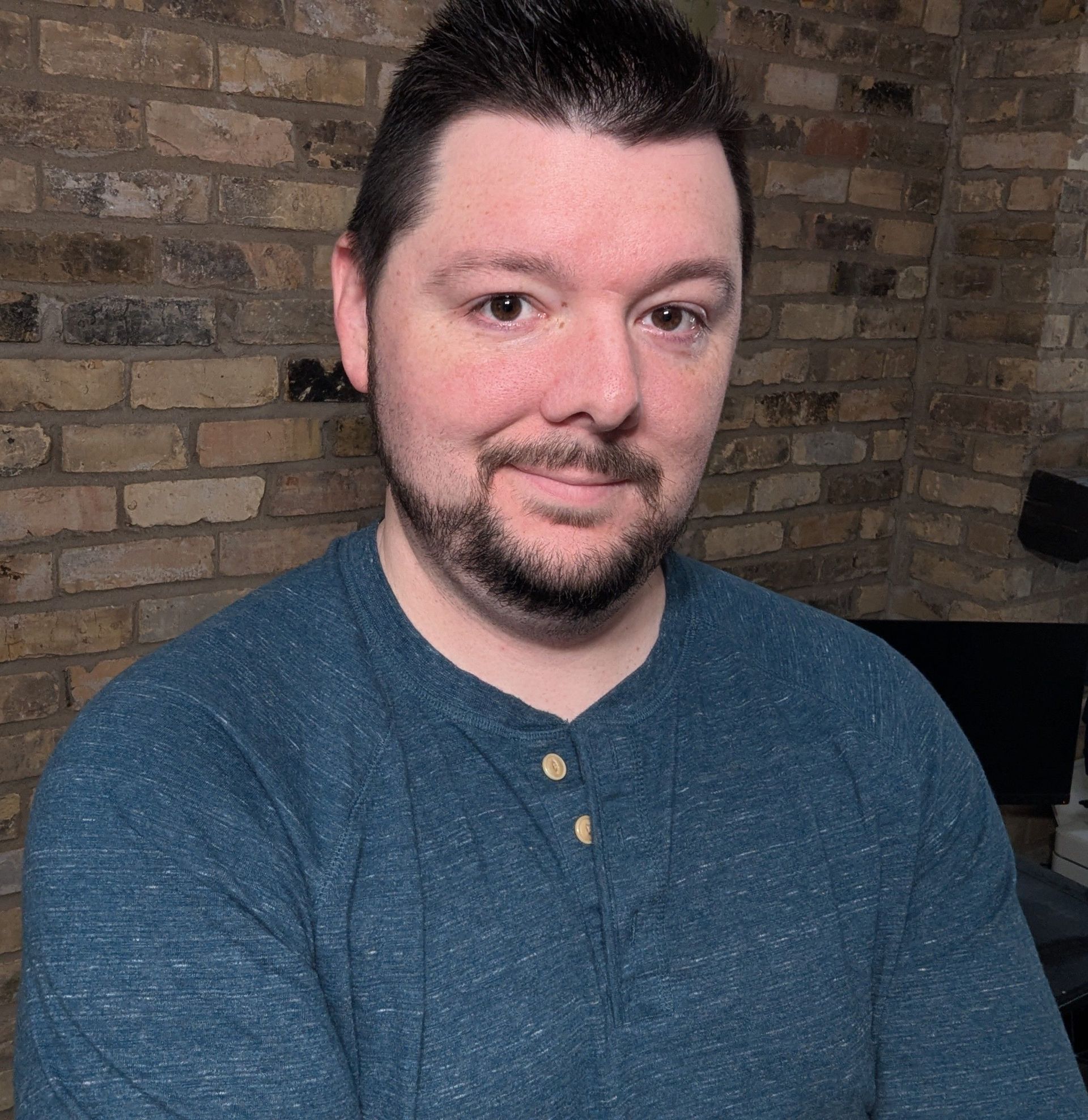 Man with dark hair and beard, wearing a blue Henley shirt, smiles, in front of a brick wall.