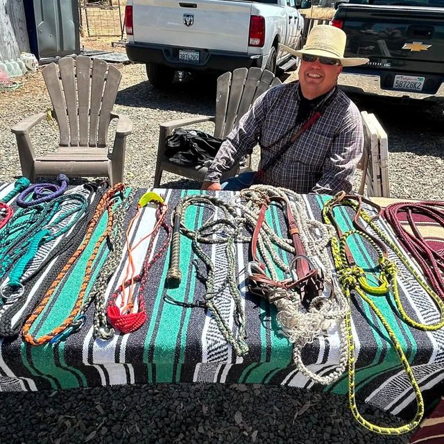 Vendor seated behind a table of colorful woven ropes and cords outdoors beside parked trucks