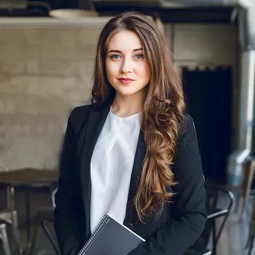 Woman in a blazer and white top, holding a notebook, in an office setting.