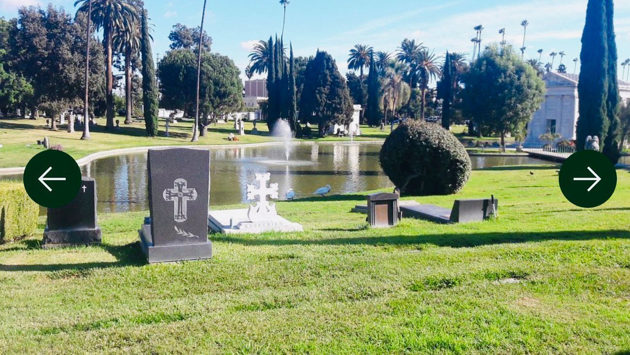 Césped verde del cementerio con árboles, lápidas y un edificio a lo largo del lado derecho bajo un cielo azul.