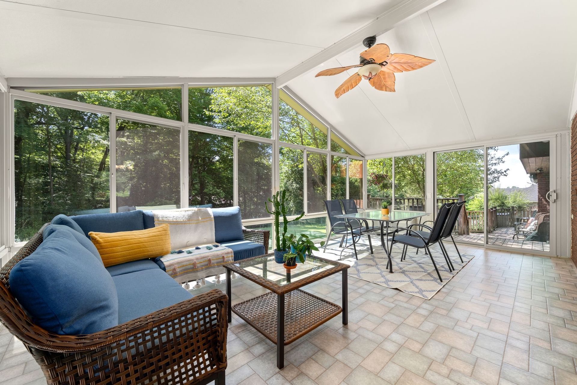 Sunroom with glass walls, wicker furniture, and a ceiling fan, overlooking a lush green yard.