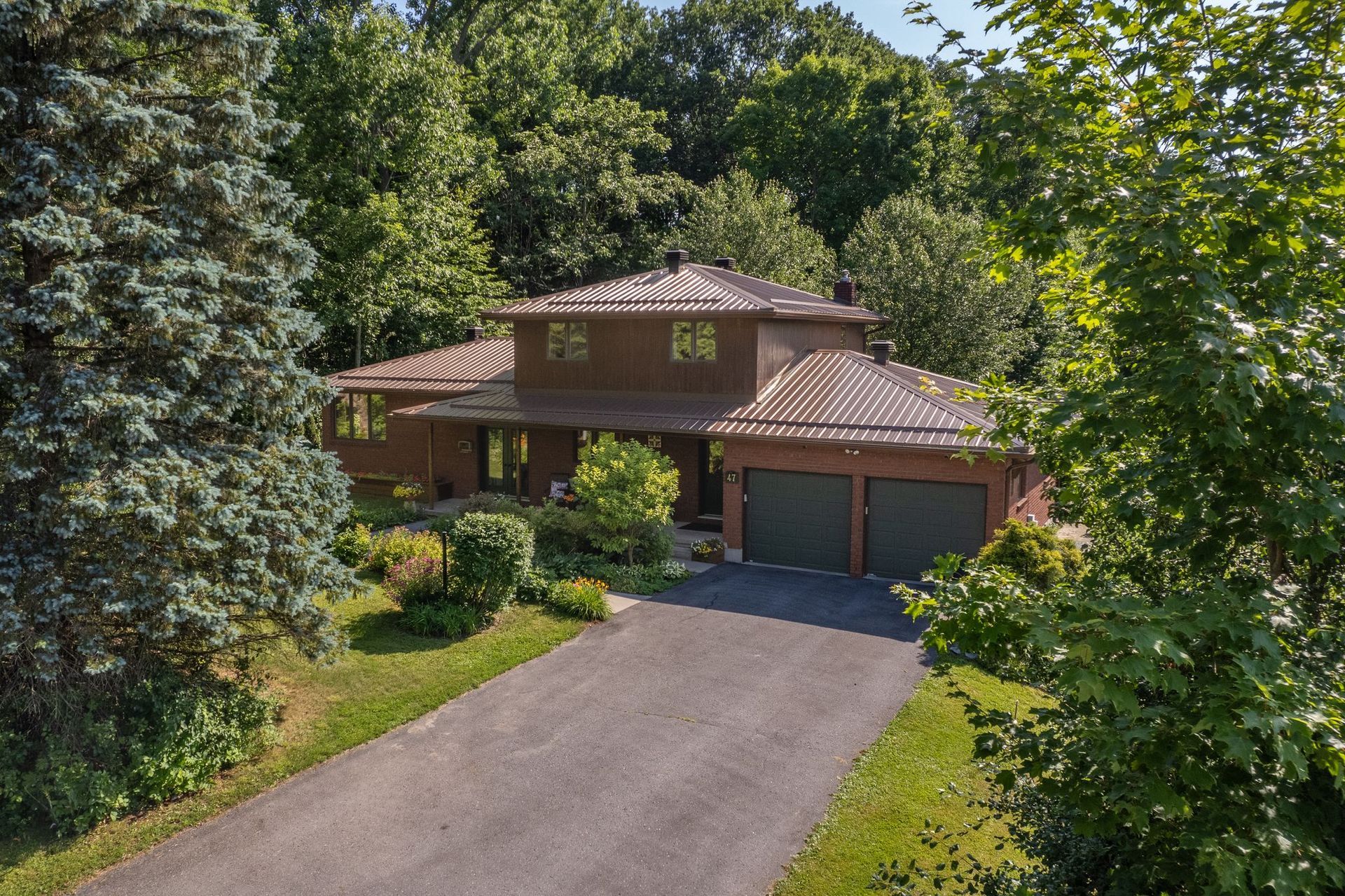 Brown house with two-car garage, driveway, and lush green trees surrounding it on a sunny day.