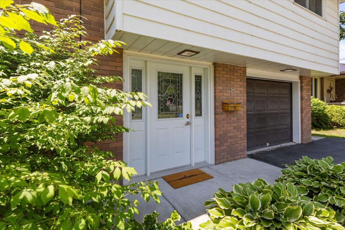 White front door with sidelights, adjacent garage. Green foliage frames the entrance.