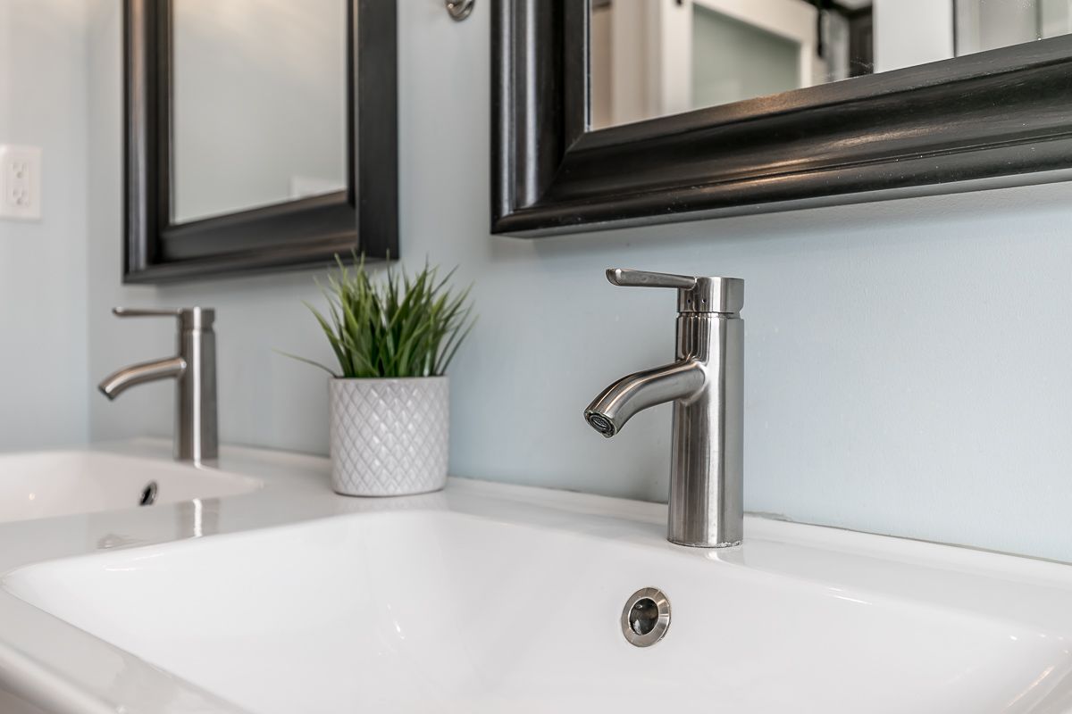 Two white sinks with modern silver faucets, green plant, and black framed mirrors on a light blue wall.