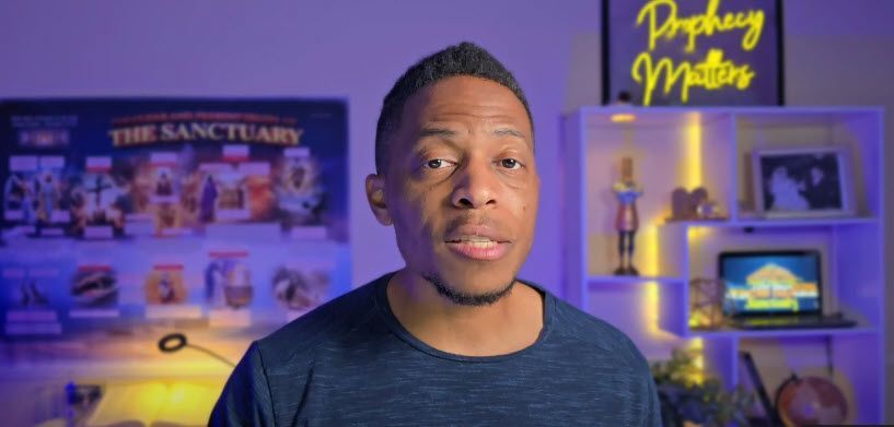 A man speaking into the camera, with a blue background, bookshelves, and a sign that says 