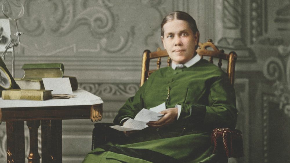 Woman in green dress seated, holding papers, beside a table with books and a clock, ornate wall in the background.