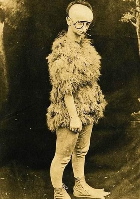 Man in fur suit, large head, wearing small glasses, standing.