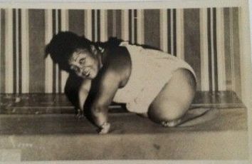Woman in a white swimsuit, posing on a bench. Striped backdrop. Leaning forward, smiling.