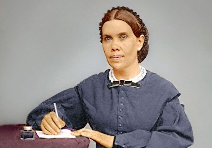 Woman in blue dress writing at a desk, looking forward.