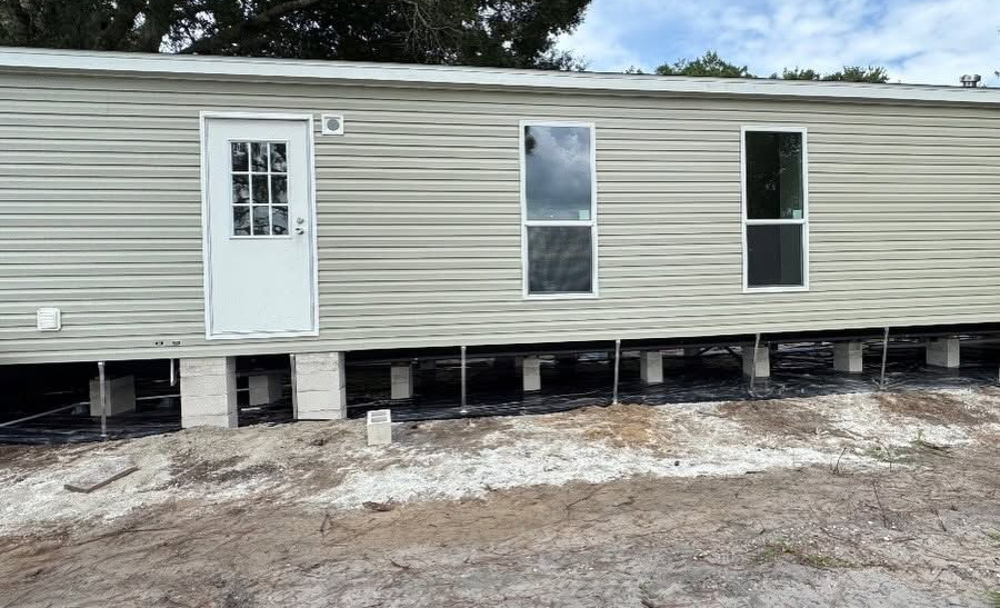 Raised beige mobile home with white door and windows, sitting on cinder blocks in a bare dirt lot.