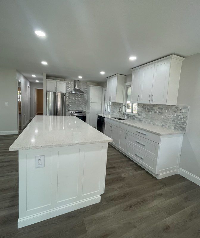 Bright modern kitchen with white cabinets, island, stainless appliances, and gray wood flooring.