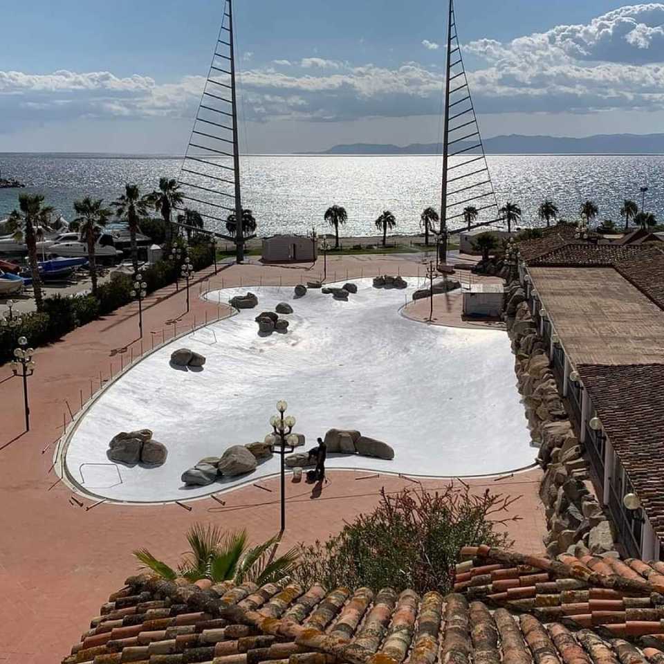 Piazza vuota con fontana bianca affacciata su un oceano luminoso, con palme e sculture a forma di vela.
