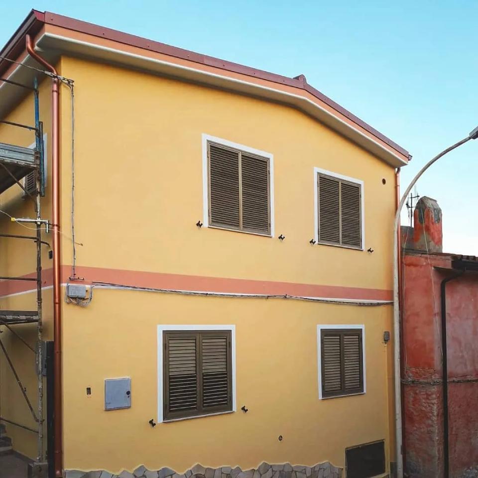 Casa gialla a due piani con finestre sbarrate e tetto rosso accanto a un muro rosa