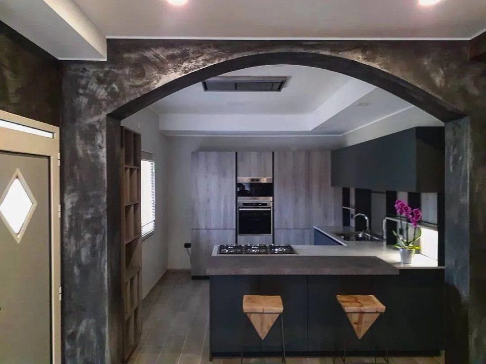 Cucina moderna vista attraverso una porta ad arco scura, con mobili grigi, un'isola e fiori viola sul bancone.