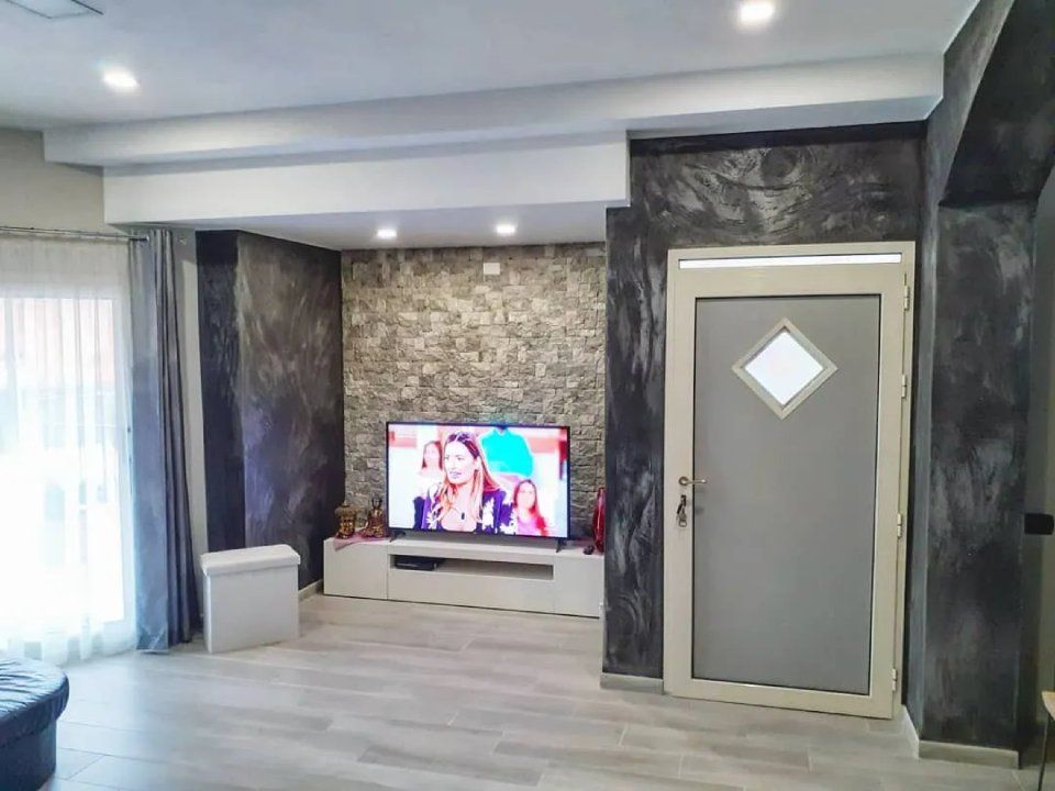 Soggiorno moderno con TV, parete decorativa piastrellata e porta grigia con finestra a rombo.