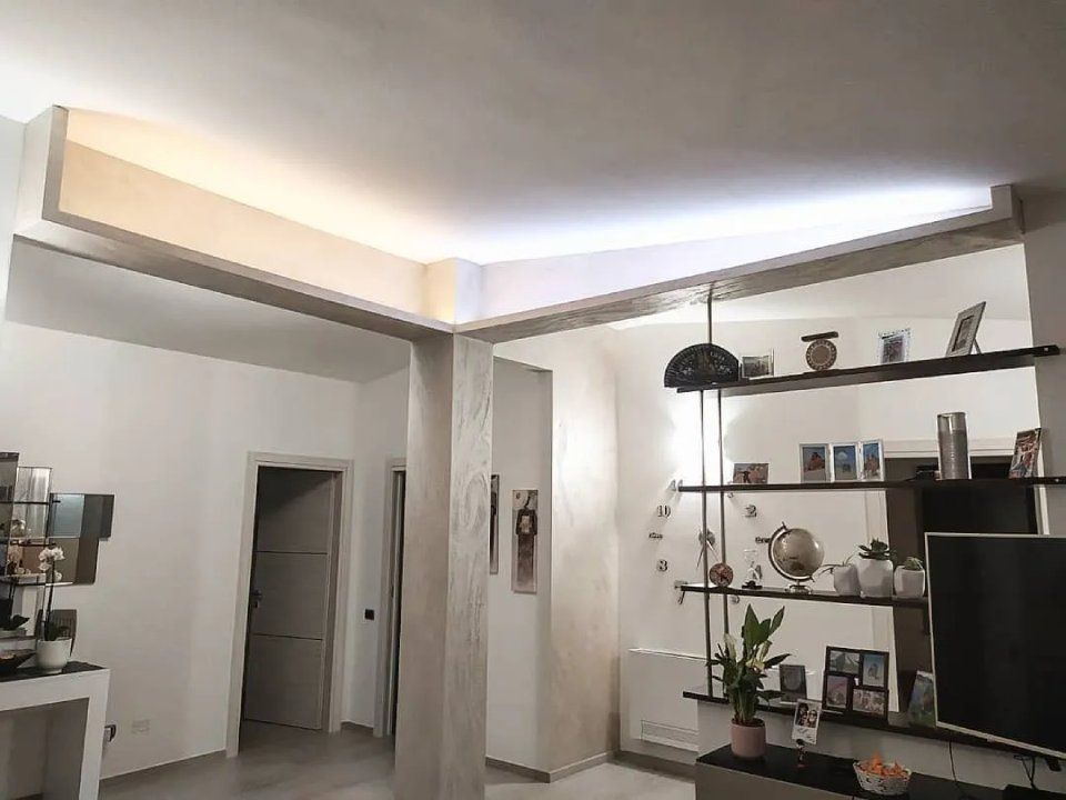 Soggiorno moderno con TV, mensole, piante e una porta illuminata da una luce soffusa proveniente dal soffitto.