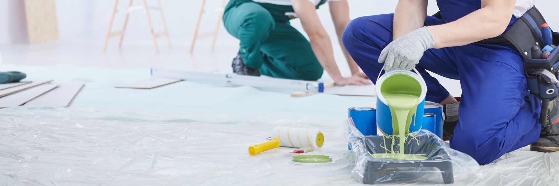 Una persona versa vernice verde da un secchio mentre è inginocchiata su un pavimento bianco durante dei lavori di tinteggiatura in casa.