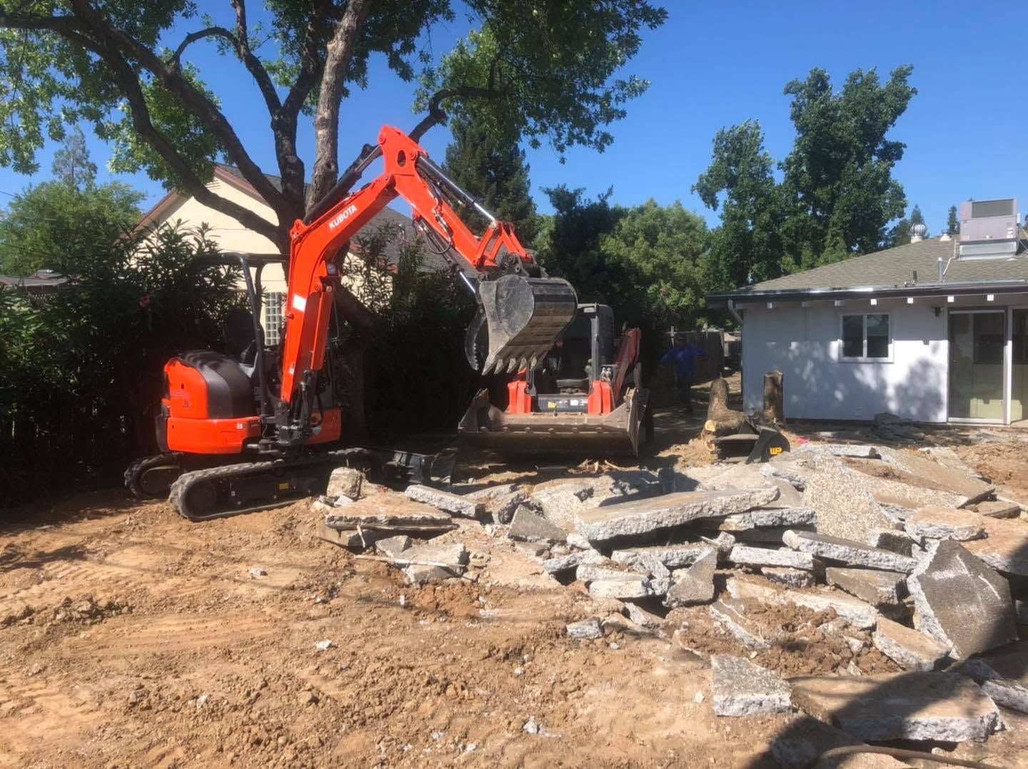 Orange excavator clearing rubble in a yard beside a white house and trees