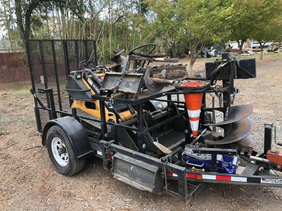 Trailer carrying a stripped-down black vehicle frame with orange cone on a dirt roadside.