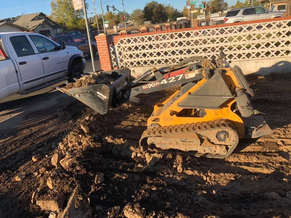 Yellow skid steer in muddy yard near a cinderblock wall, with a pickup truck and debris nearby