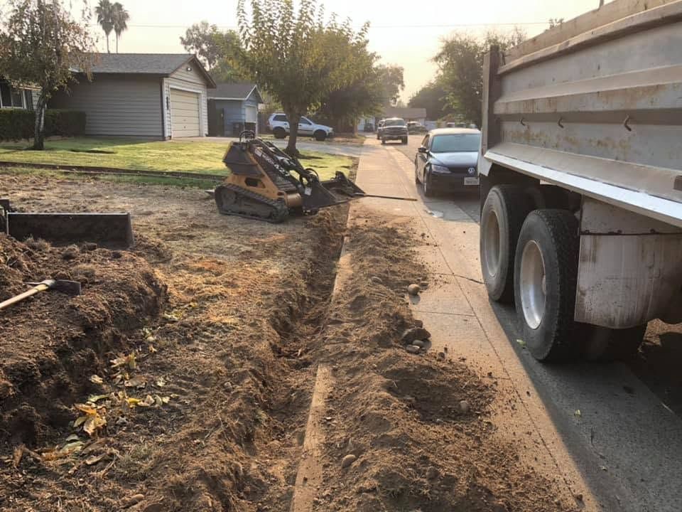 Construction truck beside a trench along a residential street, with a backhoe and parked cars nearby