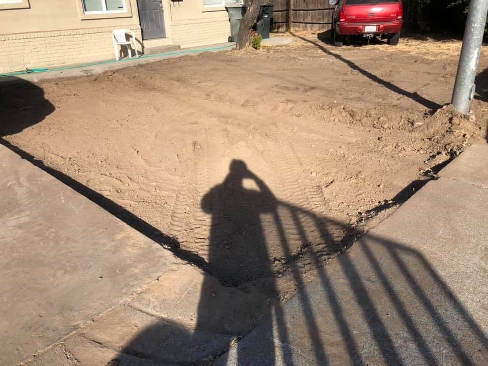 Shallow dirt-filled yard beside a house and red car, with a person’s shadow and fence shadow across the ground