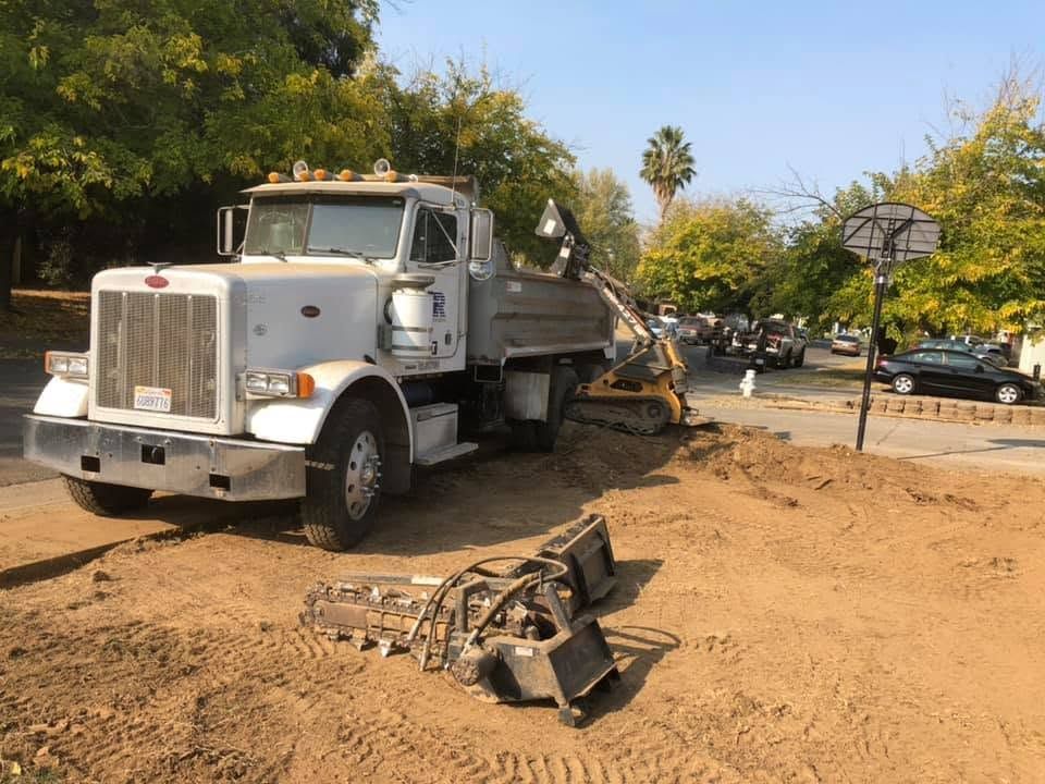 White dump truck and excavator on a dirt construction site near trees and parked cars