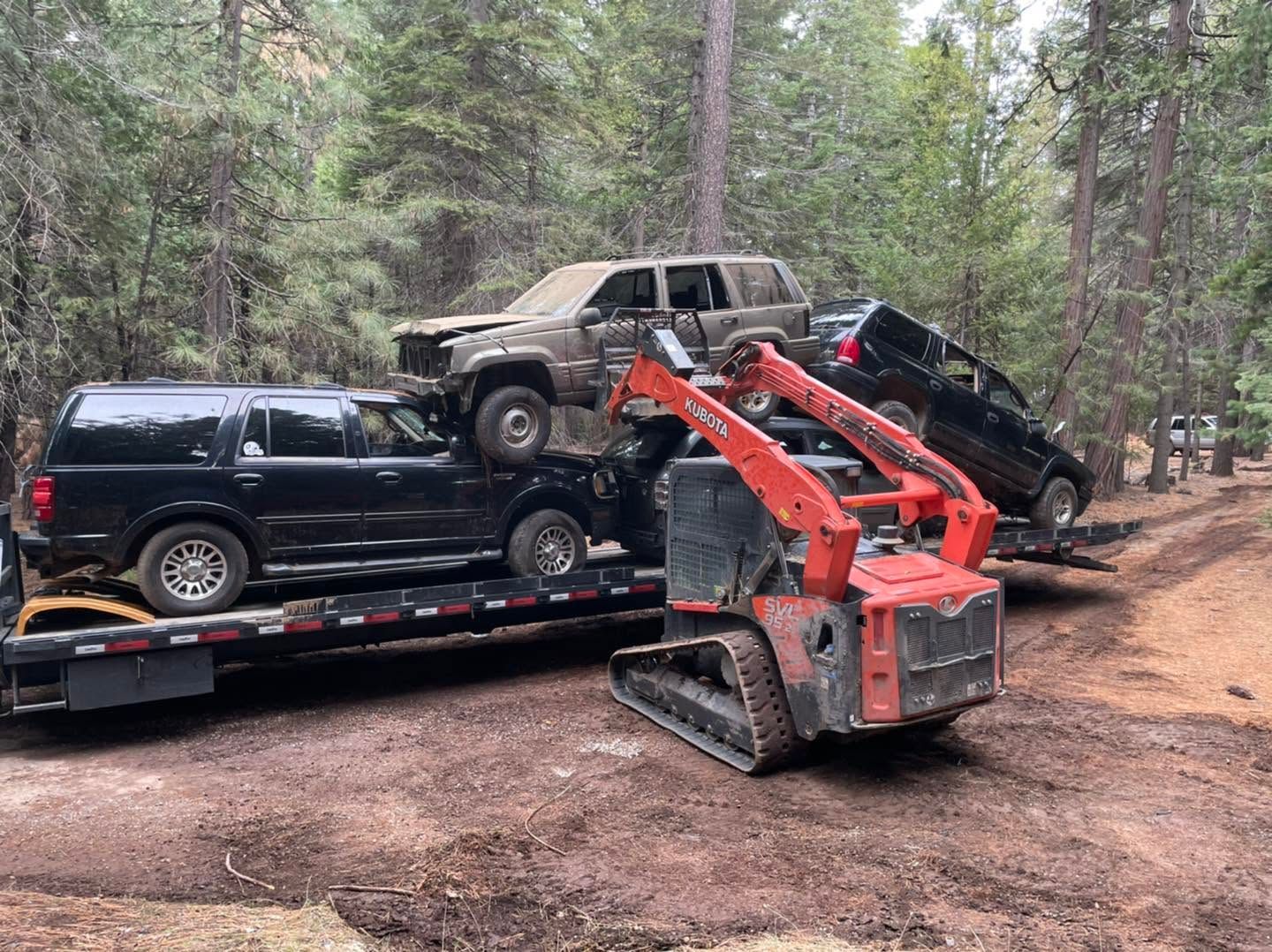 Three damaged SUVs and a red mini excavator on a trailer in a wooded area