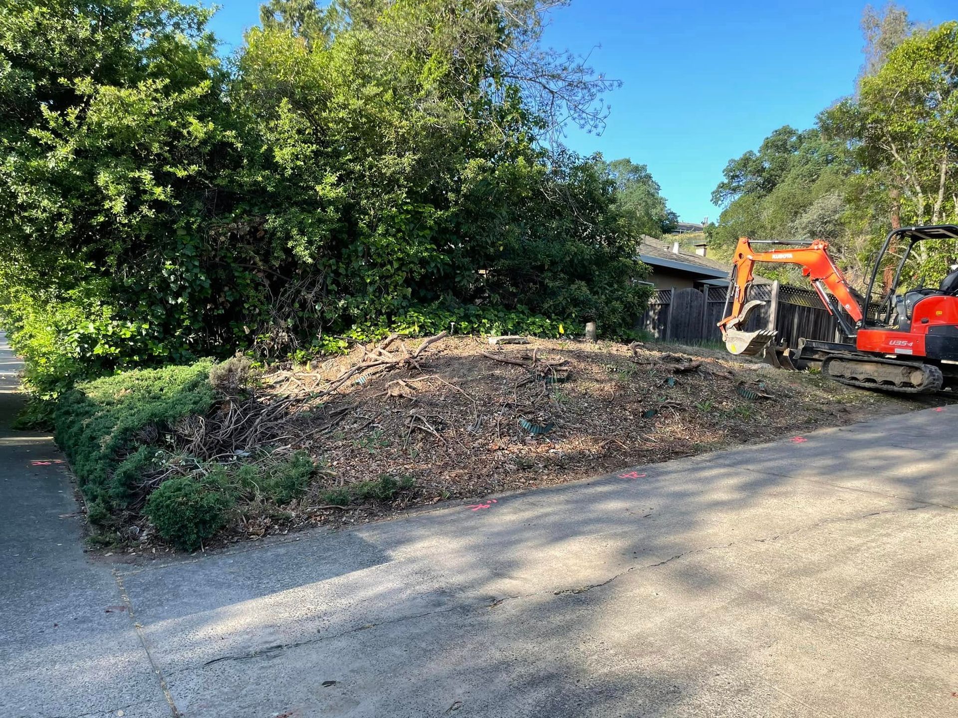 Construction equipment clearing a large dirt pile beside a road, with dense green trees and blue sky behind.