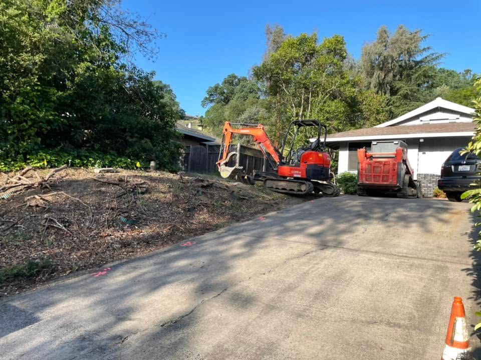Driveway with an excavator and red dump truck beside a house, clearing a wooded lot.