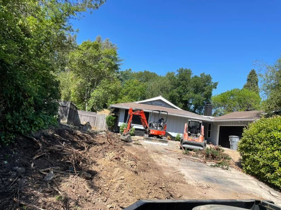 Driveway under construction with an excavator in front of a house, surrounded by trees.