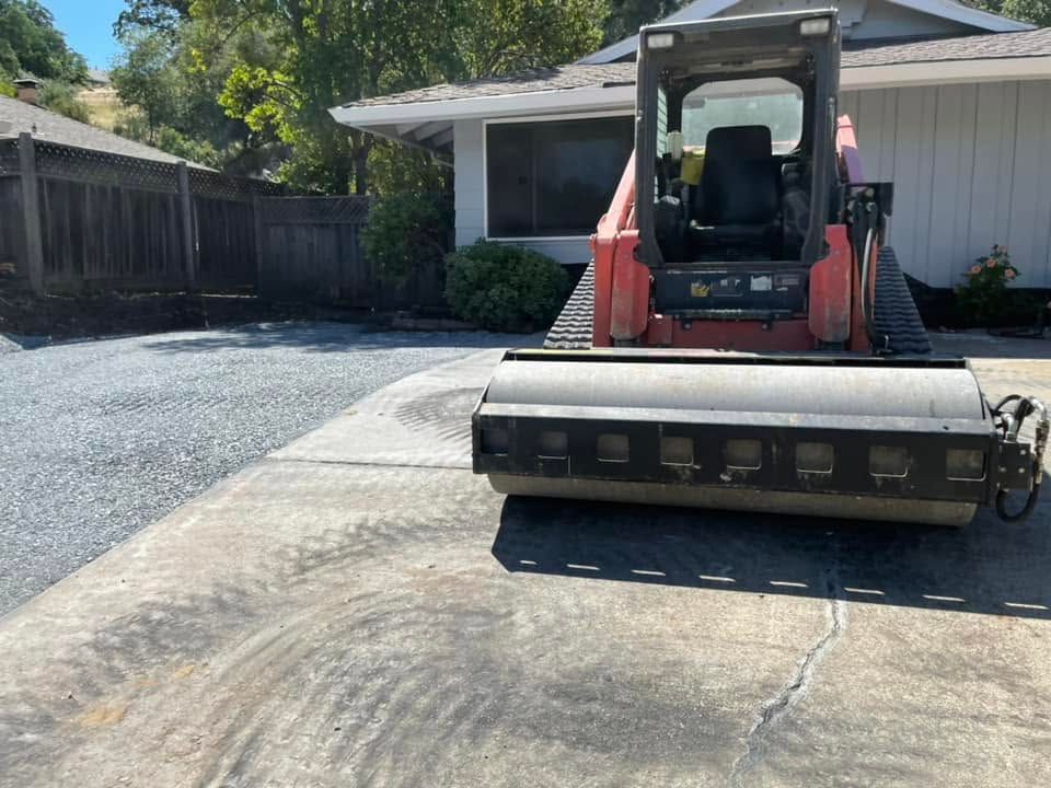 Compact excavator parked on a concrete driveway beside a white house and gravel yard
