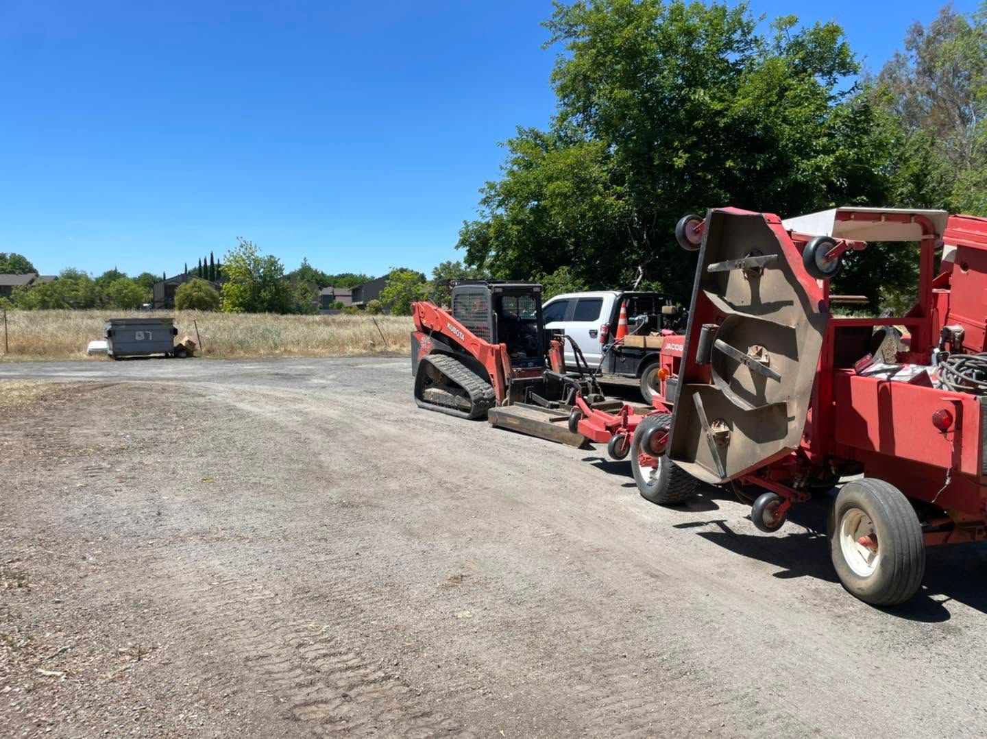 Red farm machinery parked on a gravel lot under a bright blue sky, with trees in the background