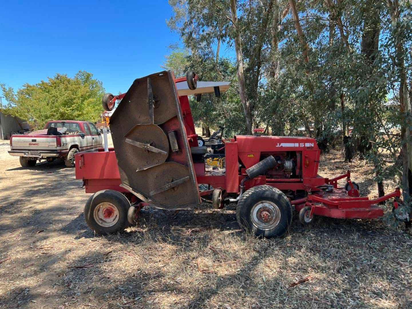 Red tractor with front mower attachment parked on dry ground near trees, with a pickup truck in the background