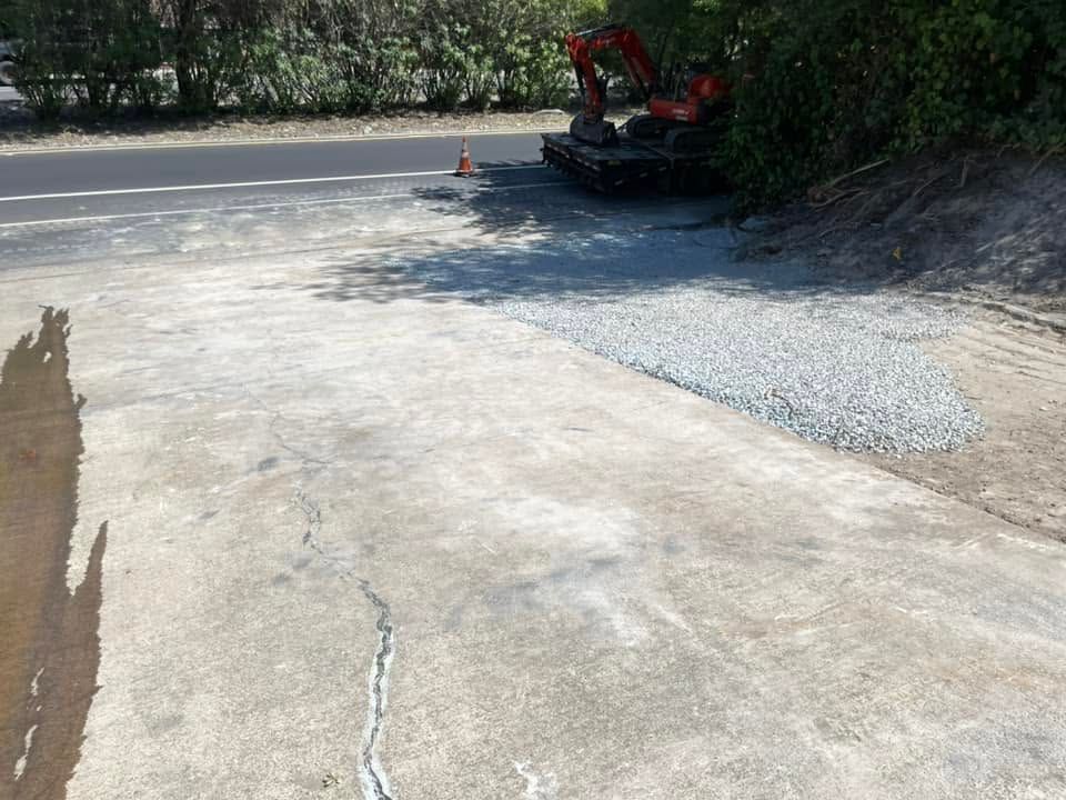 Excavator by a riverbank beside a freshly poured concrete path in a wooded area