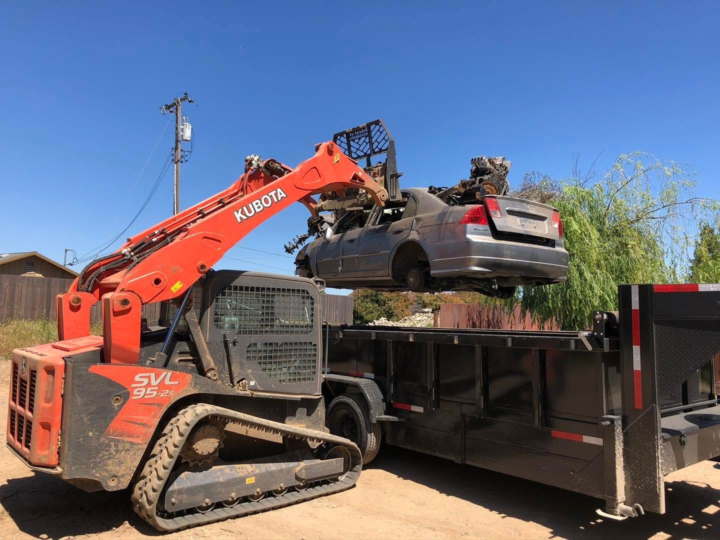 Orange excavator loading debris into a black trailer outdoors under a blue sky