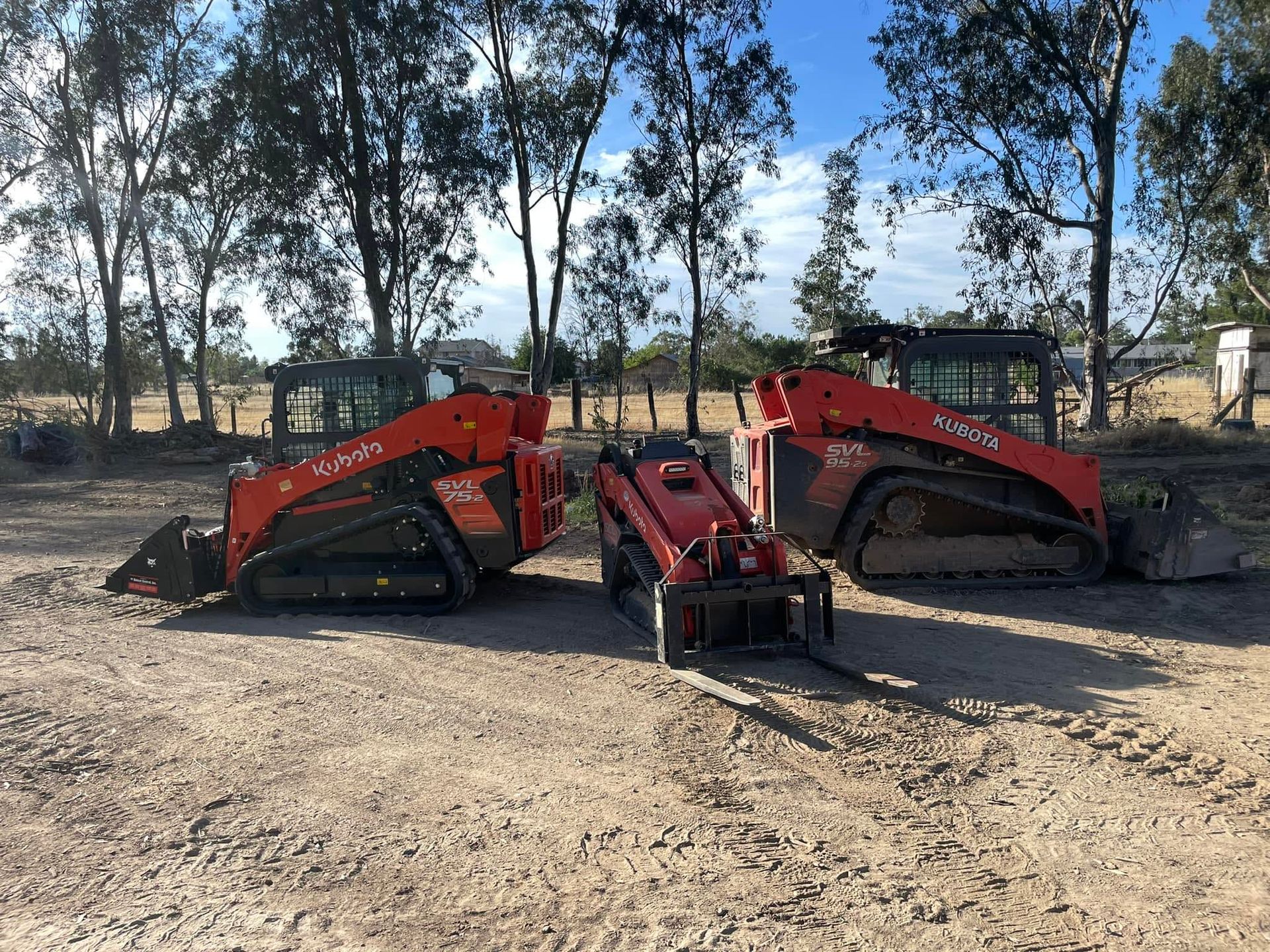 Two red skid-steer loaders parked side by side on a dirt lot with trees in the background