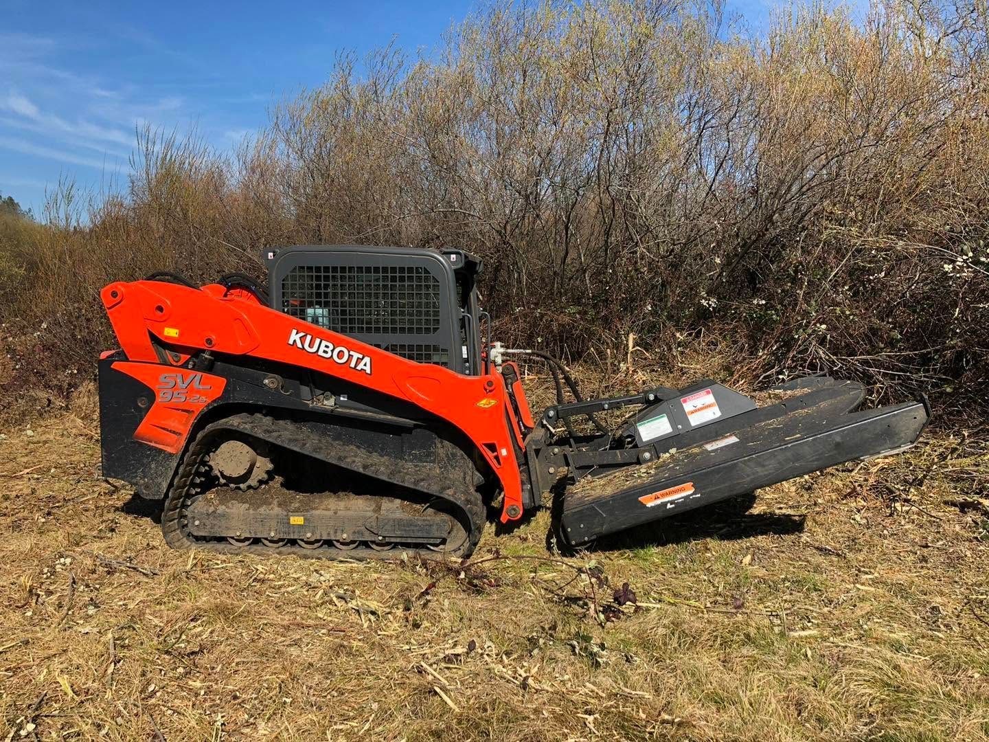 Orange Kubota skid steer with front attachment parked on a dirt path beside bushes under a blue sky