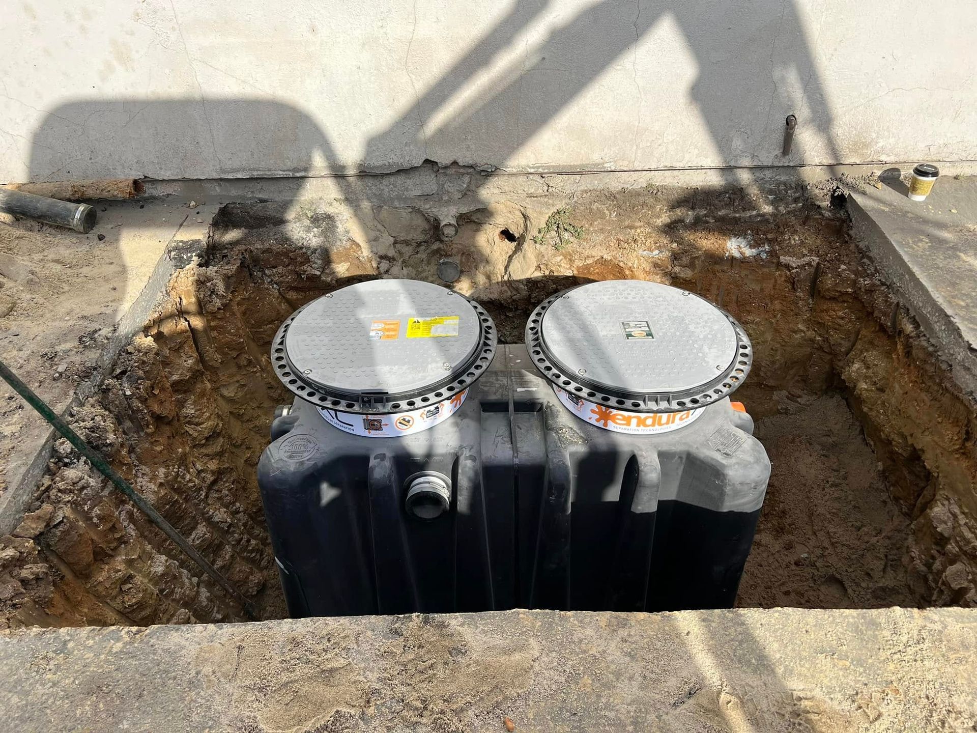 Two black plastic tanks with silver lids installed in a dirt excavation beside a concrete wall.