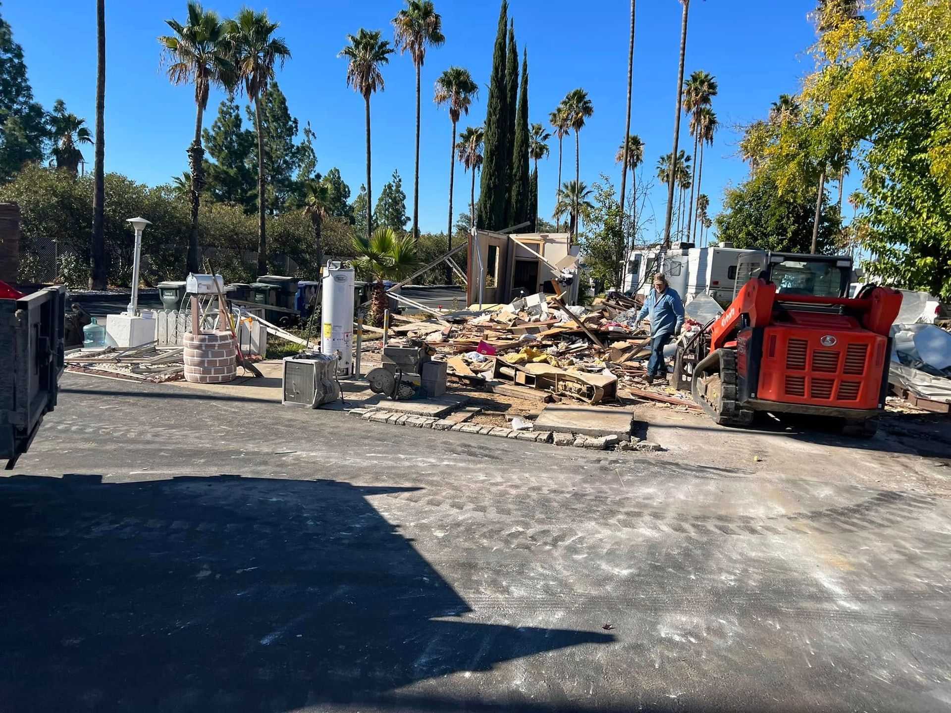Demolition debris with workers and a red dumpster in a palm-lined outdoor lot under a clear blue sky
