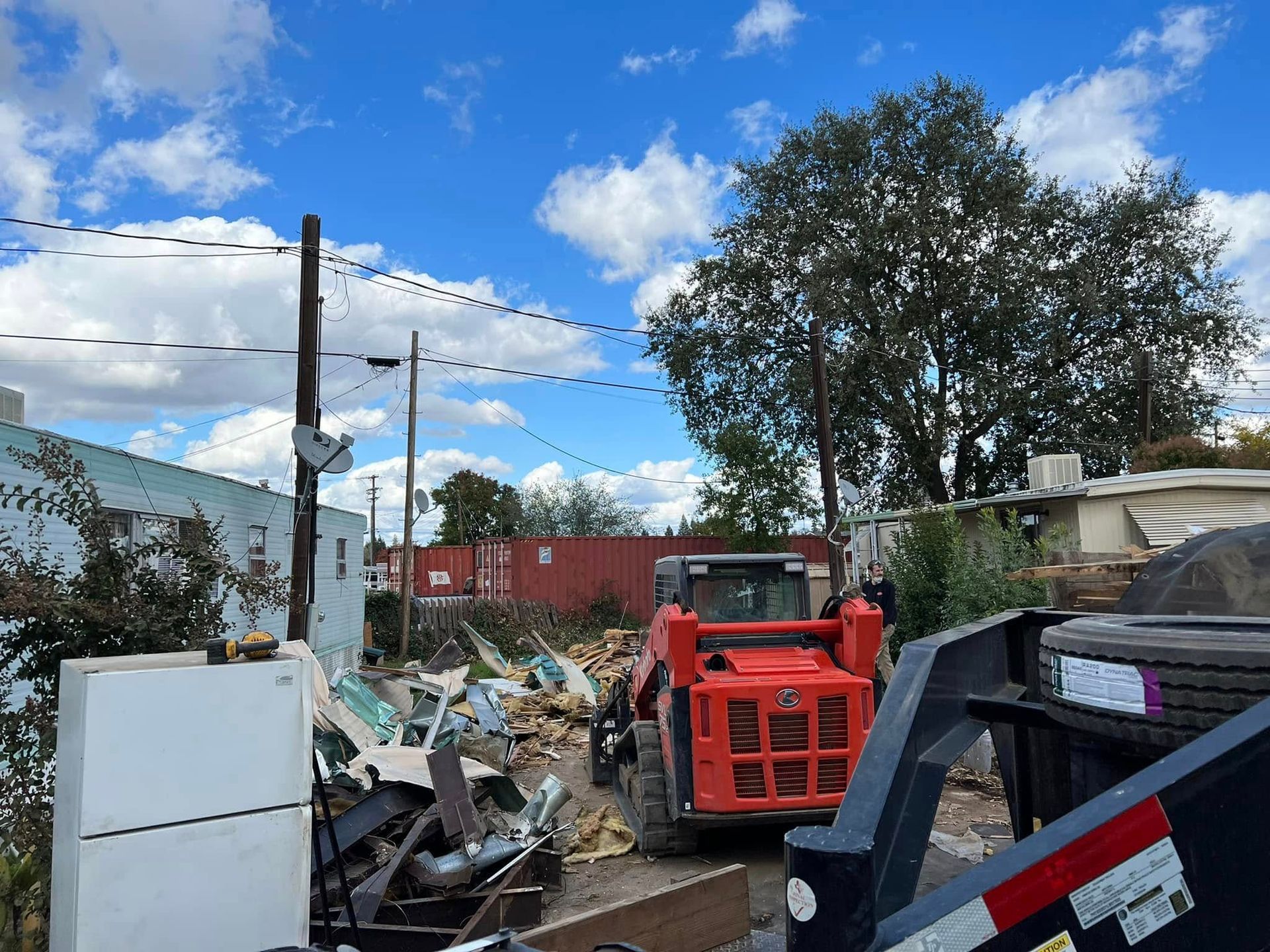 Debris-filled lot with a red dumpster, scattered scrap, utility poles, and a tree under a partly cloudy sky