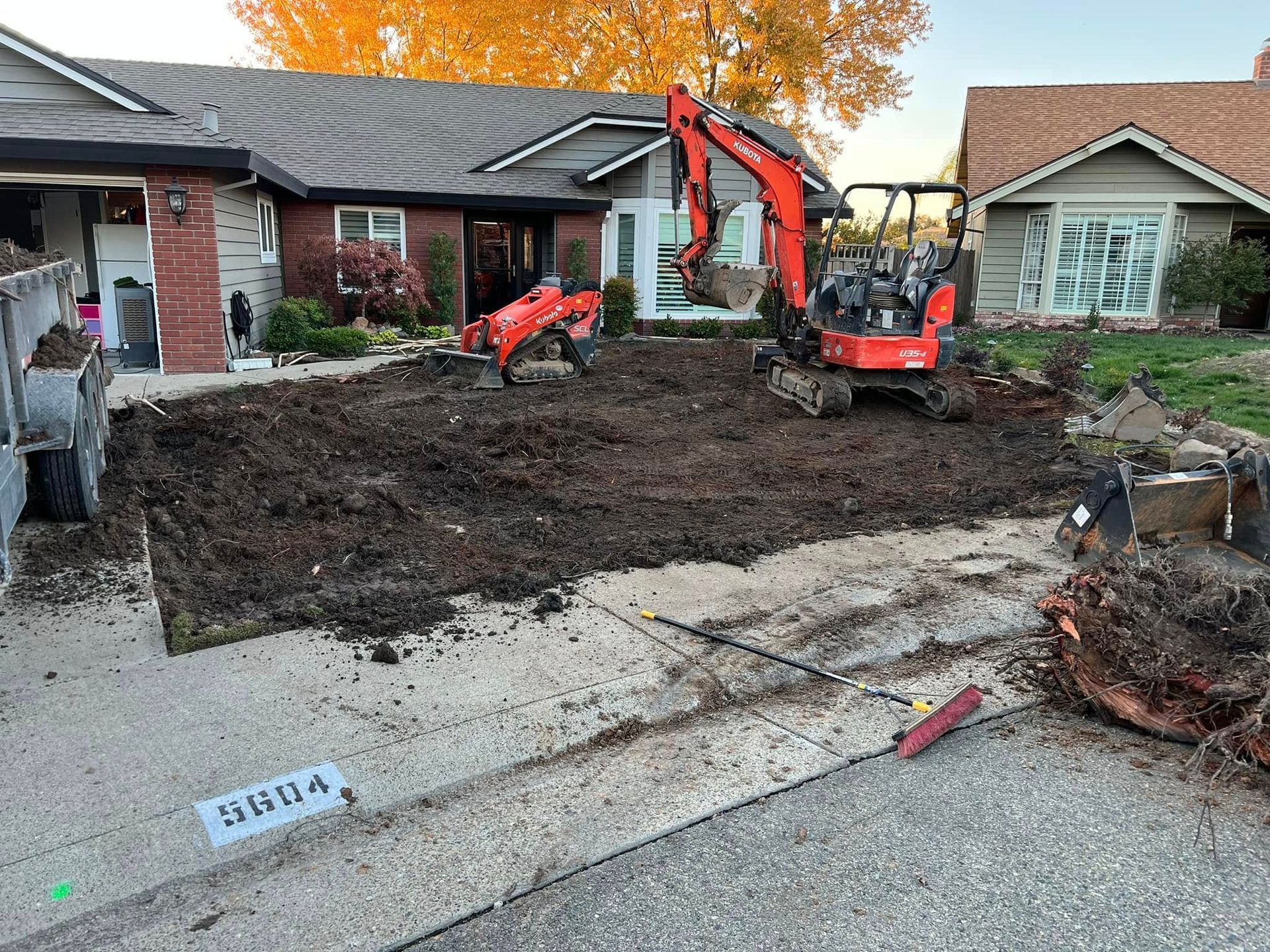Excavator and skid steer digging out a front yard beside a brick house and driveway.