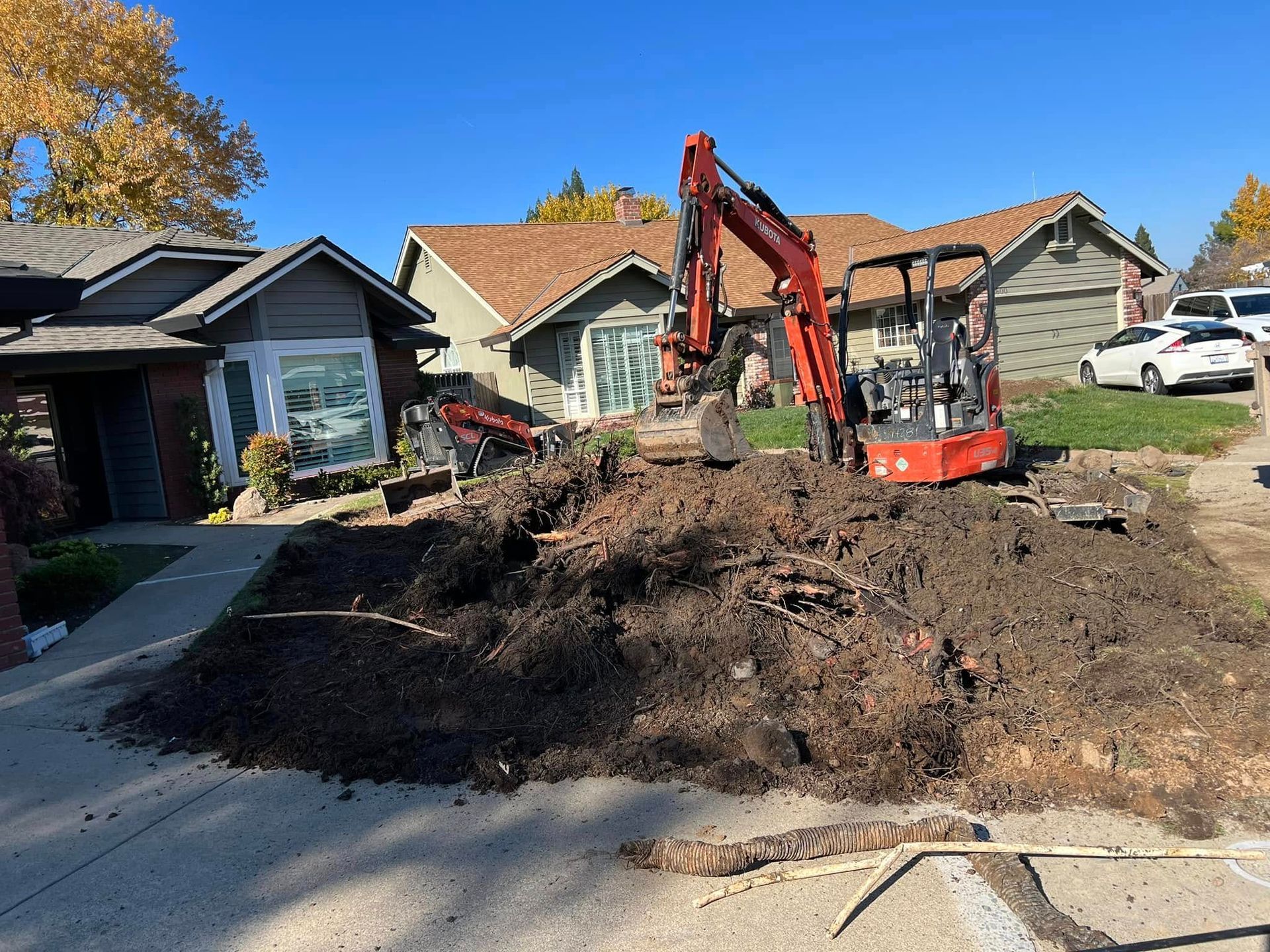 Mini excavator digging a dirt mound in a suburban driveway with houses in the background